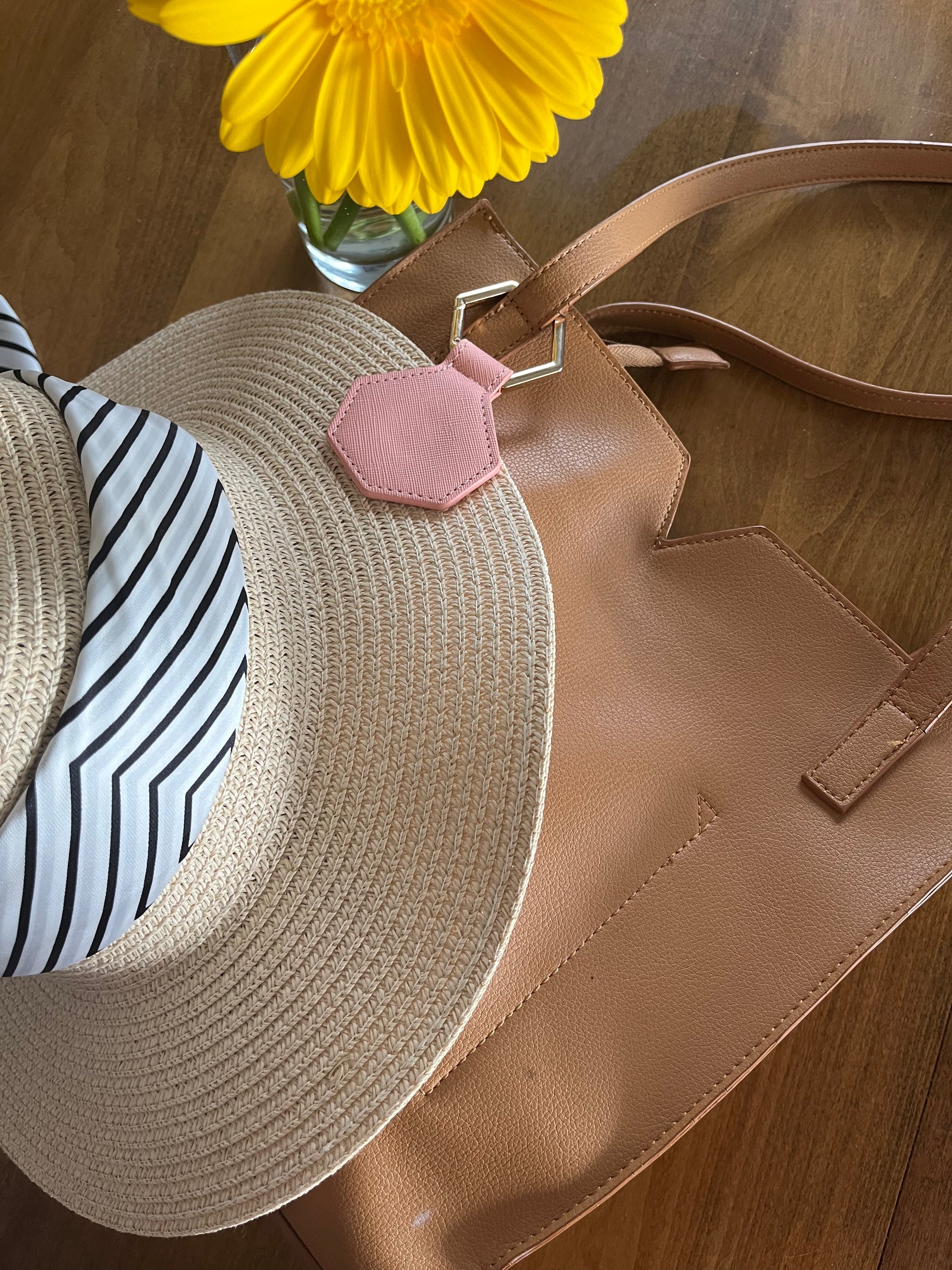 Straw hat with a striped band and pink flower accessory on a brown leather bag with a yellow flower in the background. with a pink gat clip and gold clasp 