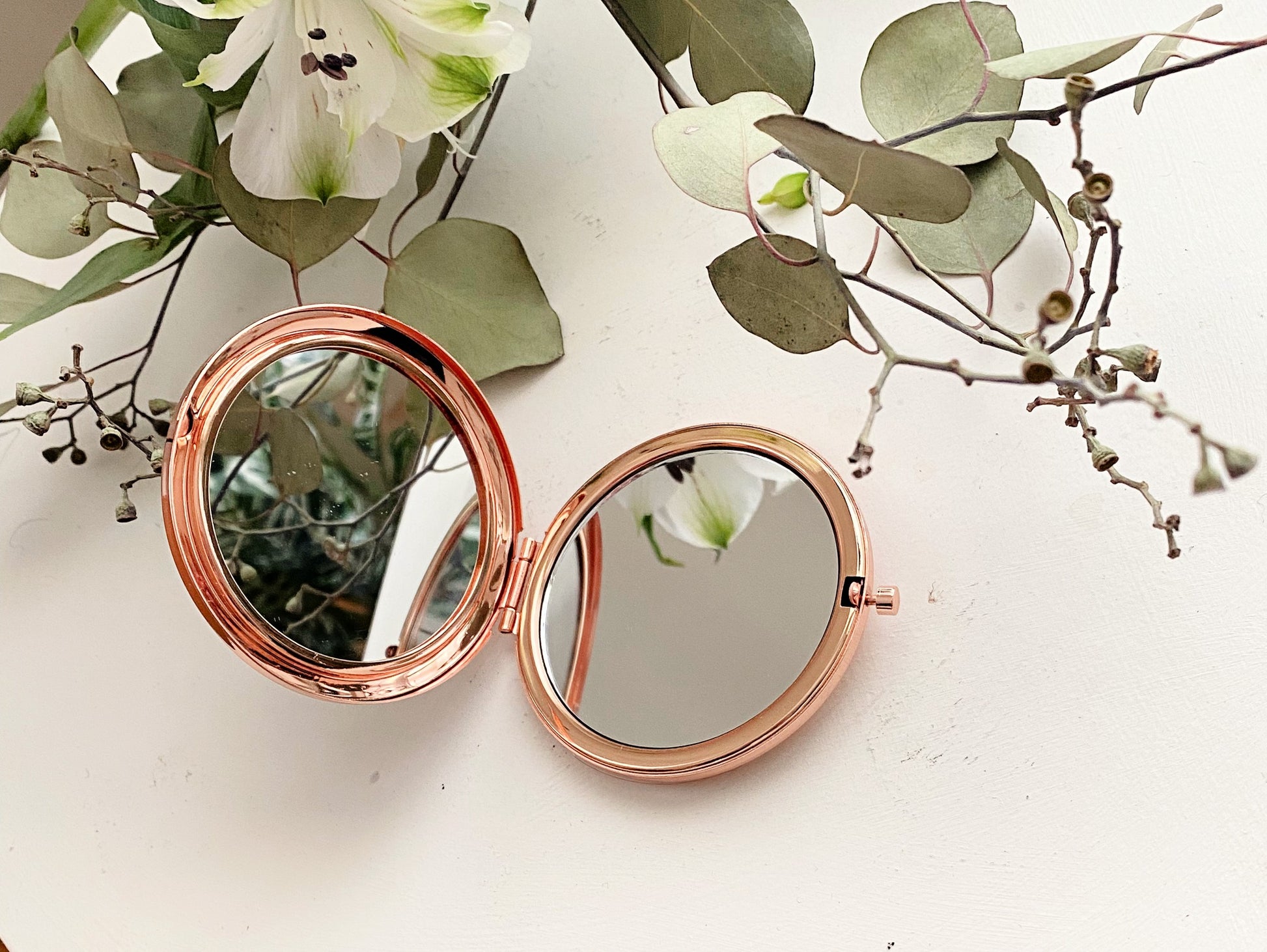 Rose gold compact mirror on a white surface with green leaves and flowers.