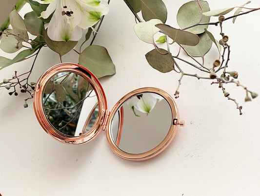 Rose gold compact mirror on a white surface with green leaves and flowers.