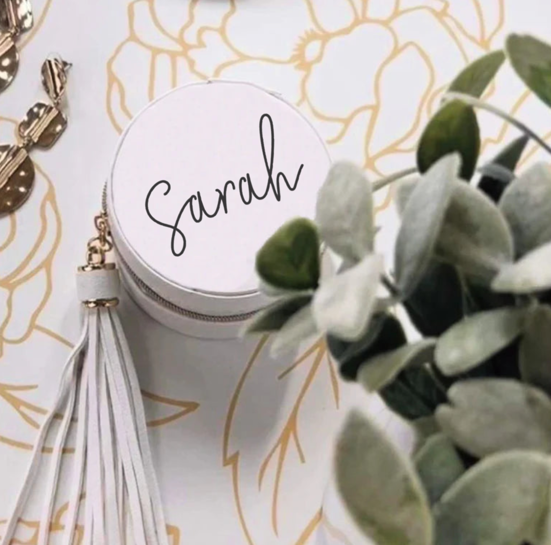 Round white container with 'Sarah' on it, tassel, and plant on a decorative background